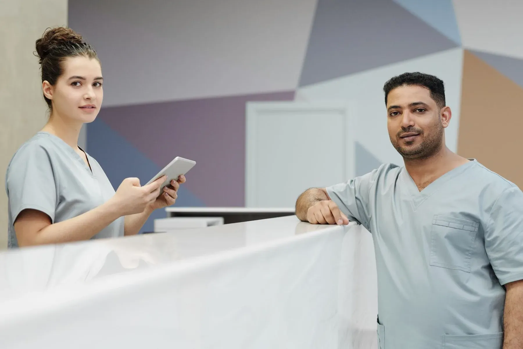 Two clinic staff in scrubs at a reception desk, one holding a tablet